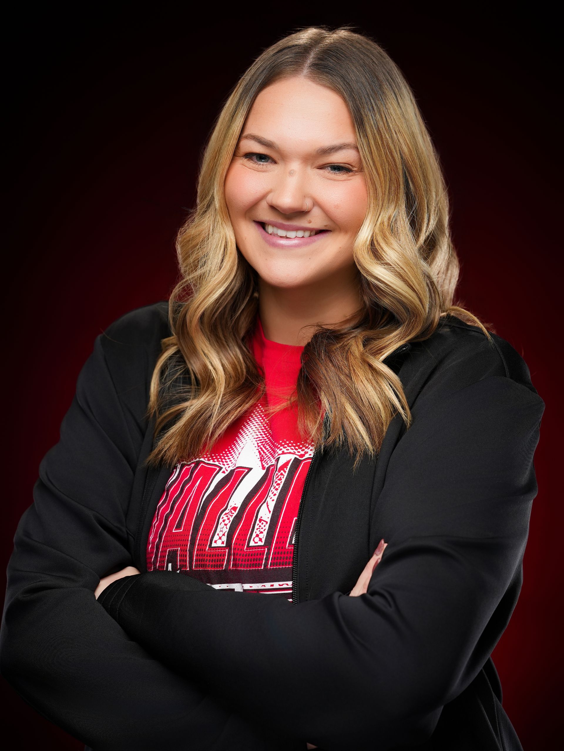 A smiling person with shoulder-length wavy hair, wearing a black jacket over a red shirt, posing with arms crossed.
