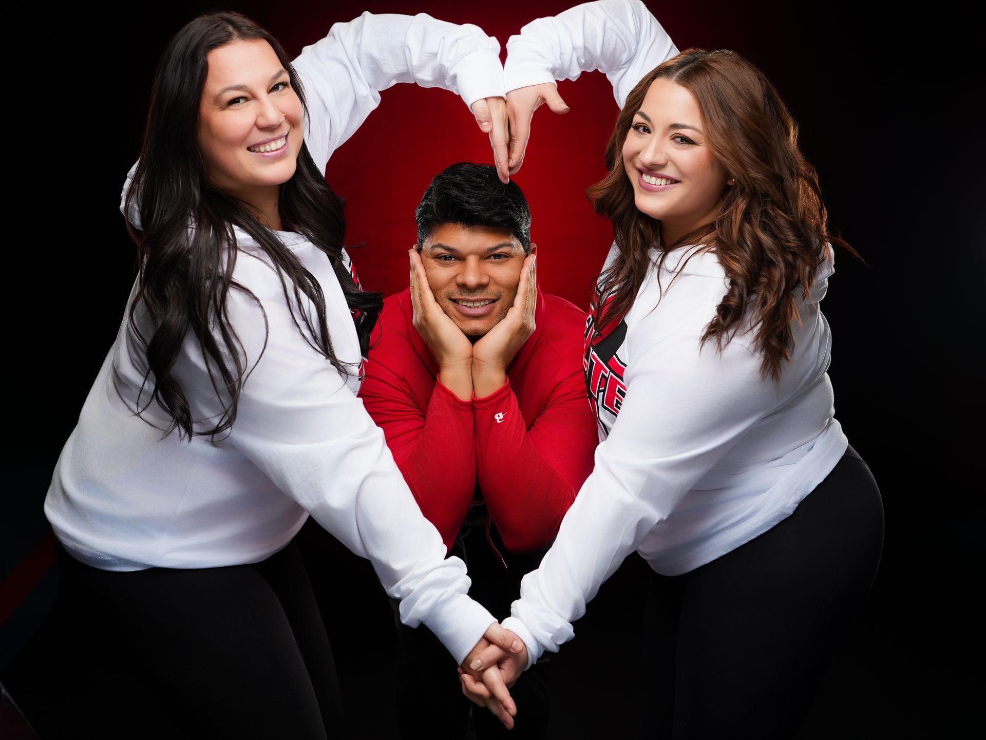 Two people in white hoodies create a heart shape with their arms around a person in a red shirt who is cupping their face.