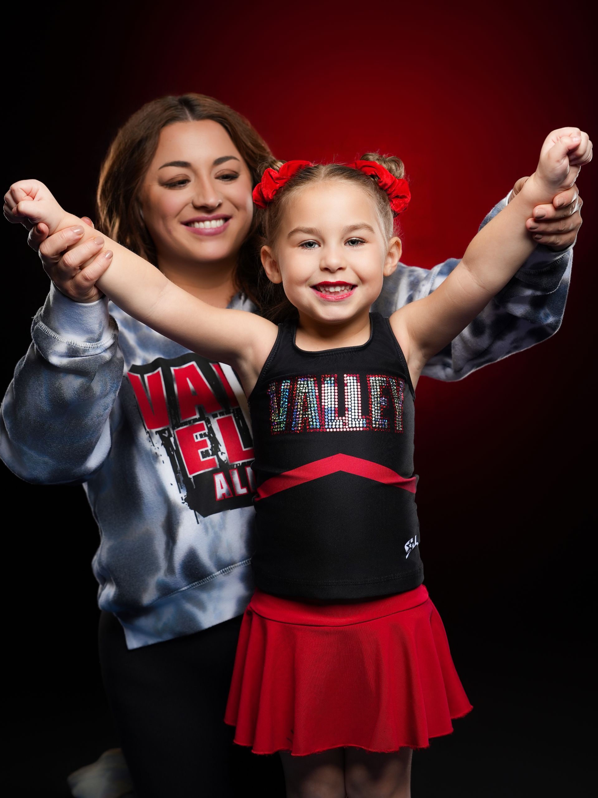 A smiling woman holding the arms of a young child in a black and red cheerleading uniform against a dark red backdrop.