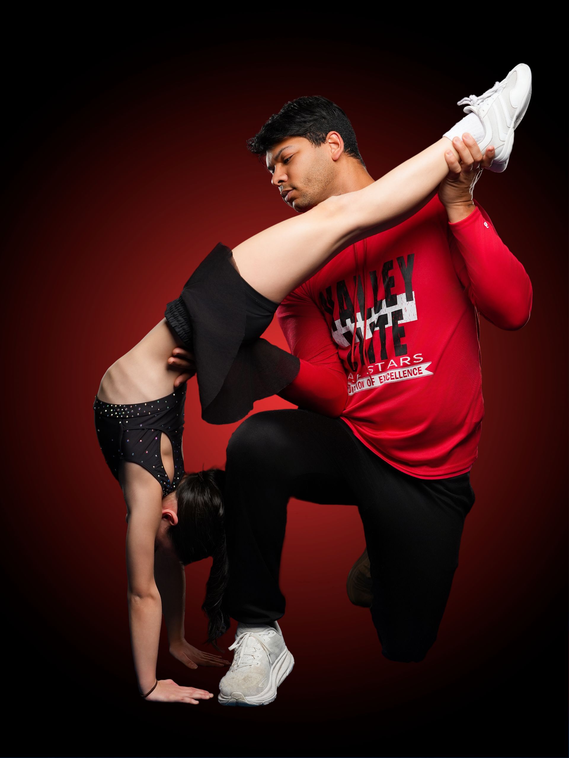 A person performs a handstand while another person supports their raised leg against a dark red gradient background.