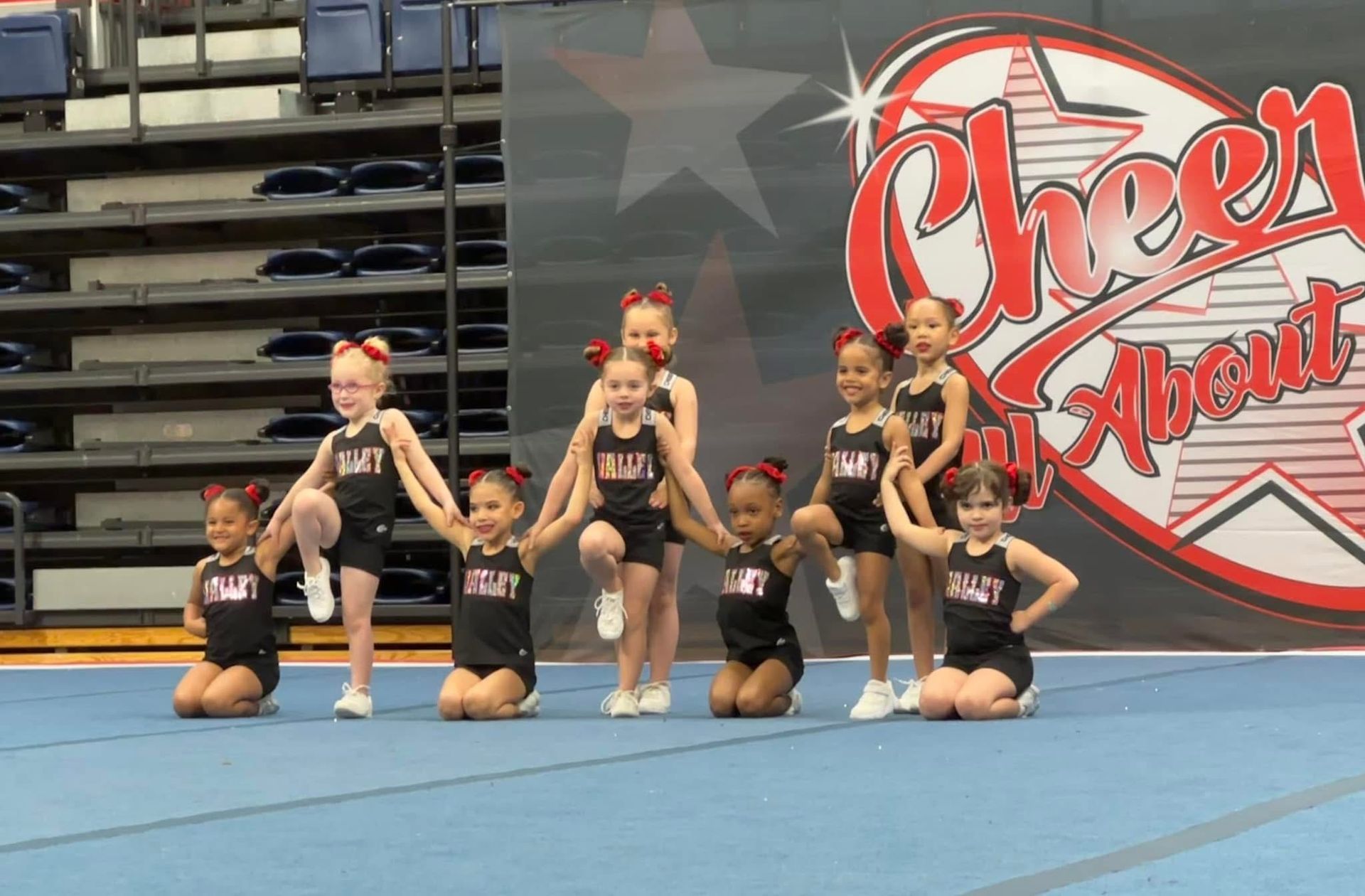A group of eight cheerleaders in black uniforms pose on a blue mat in front of a red and white branded backdrop.