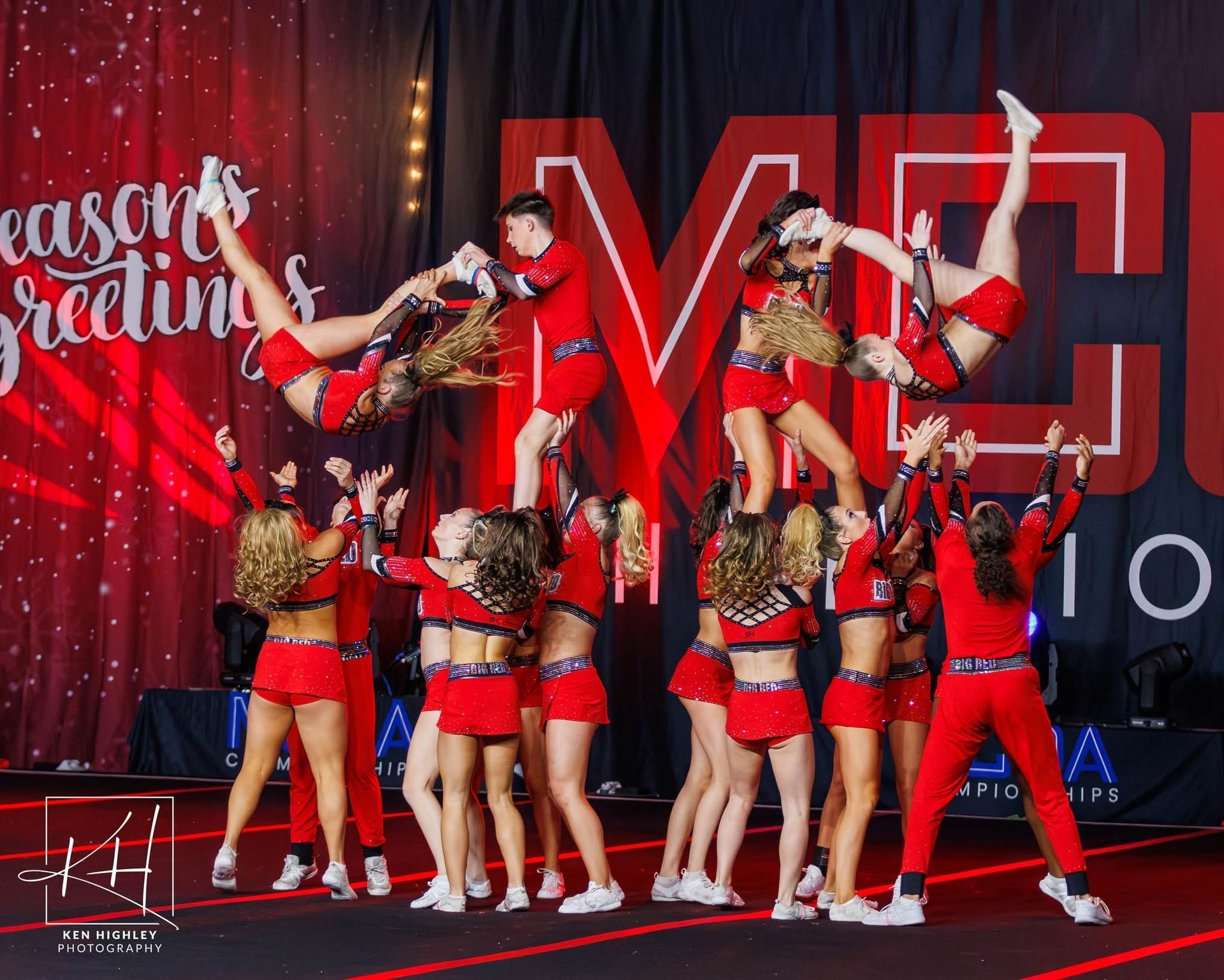 Cheerleaders in red uniforms perform a stunt with two members lifted high in the air on a stage with a holiday backdrop.