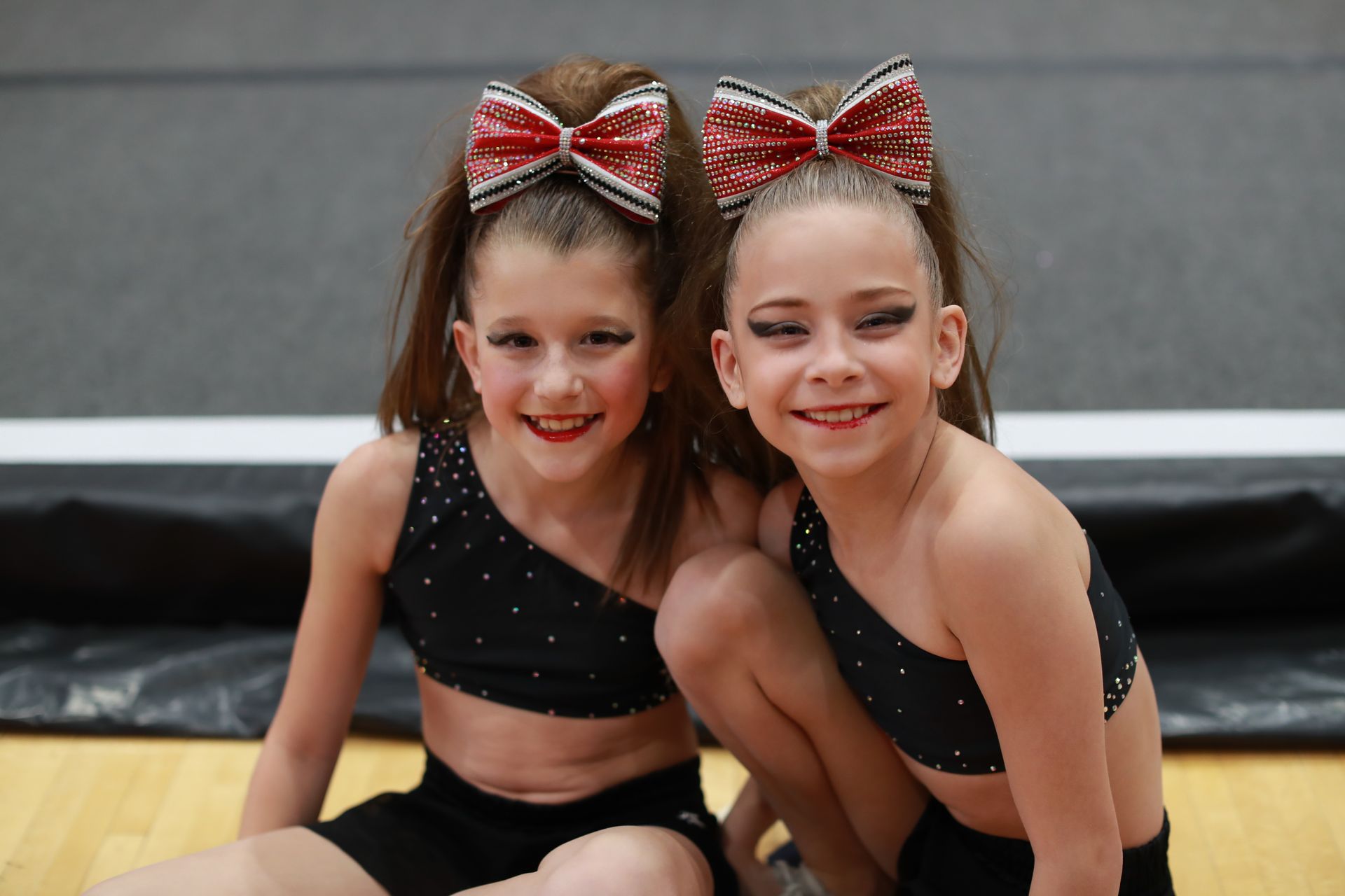 Two cheerleaders in black embellished crop tops and large red bows pose smiling on a gym floor.