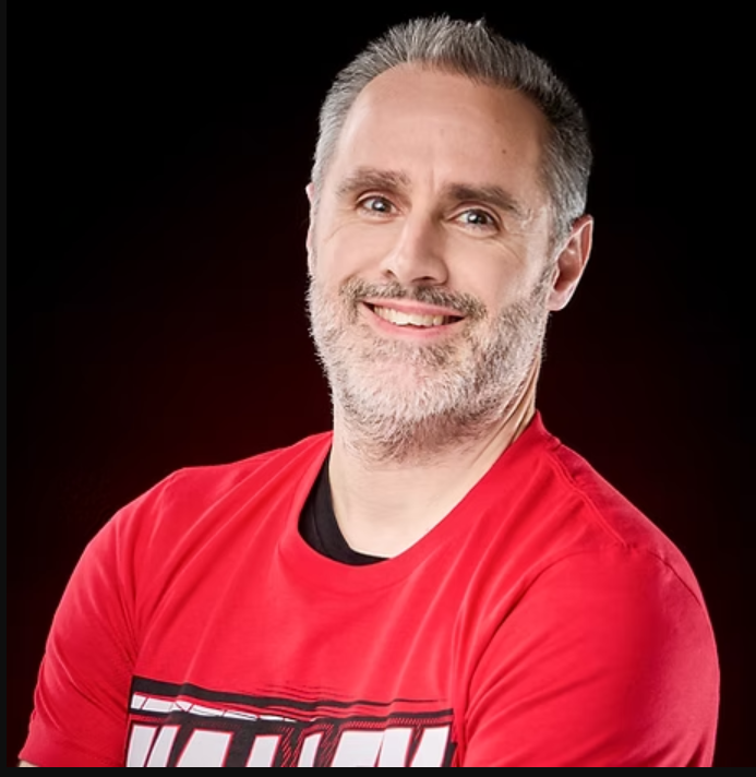 A smiling person with short, graying hair and a beard, wearing a red t-shirt against a dark background.