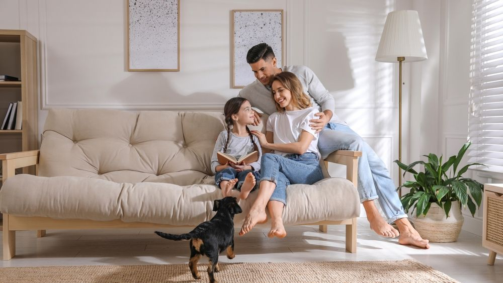 Family on couch with dog, indoor setting. Girl reading book; parents smiling, light, natural wood.