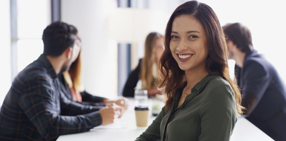 Woman smiles at the viewer in a meeting, others blurred in the background. Green shirt, bright office setting.