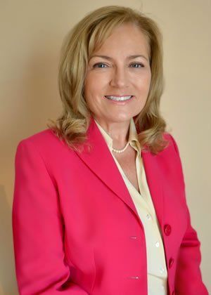 Woman in a pink blazer, smiling, standing in front of a beige background.