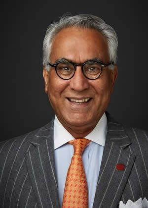Man in a pinstripe suit, glasses, and orange tie, smiling against a dark background.