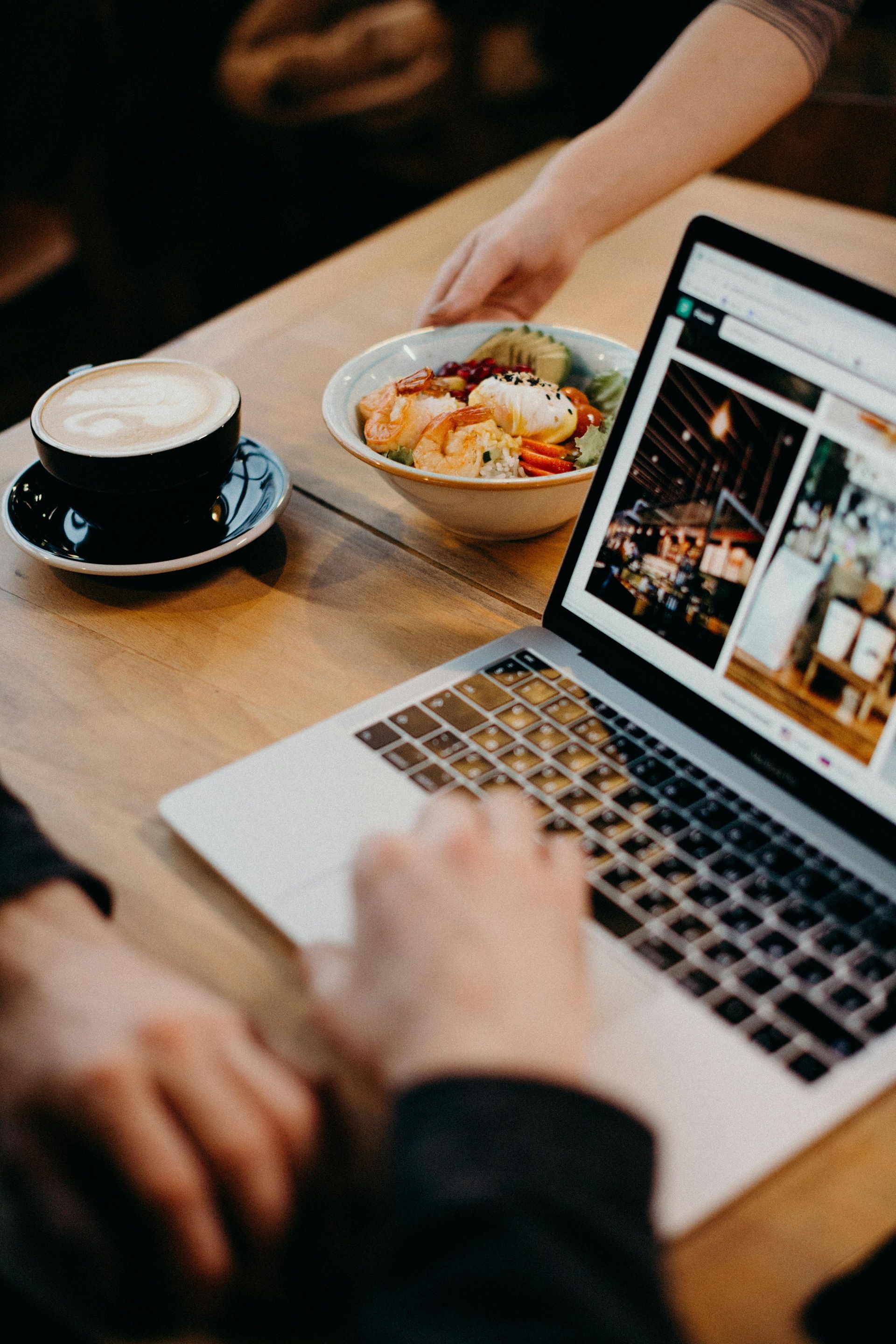 Person using laptop at table with coffee and salad; someone serving food.