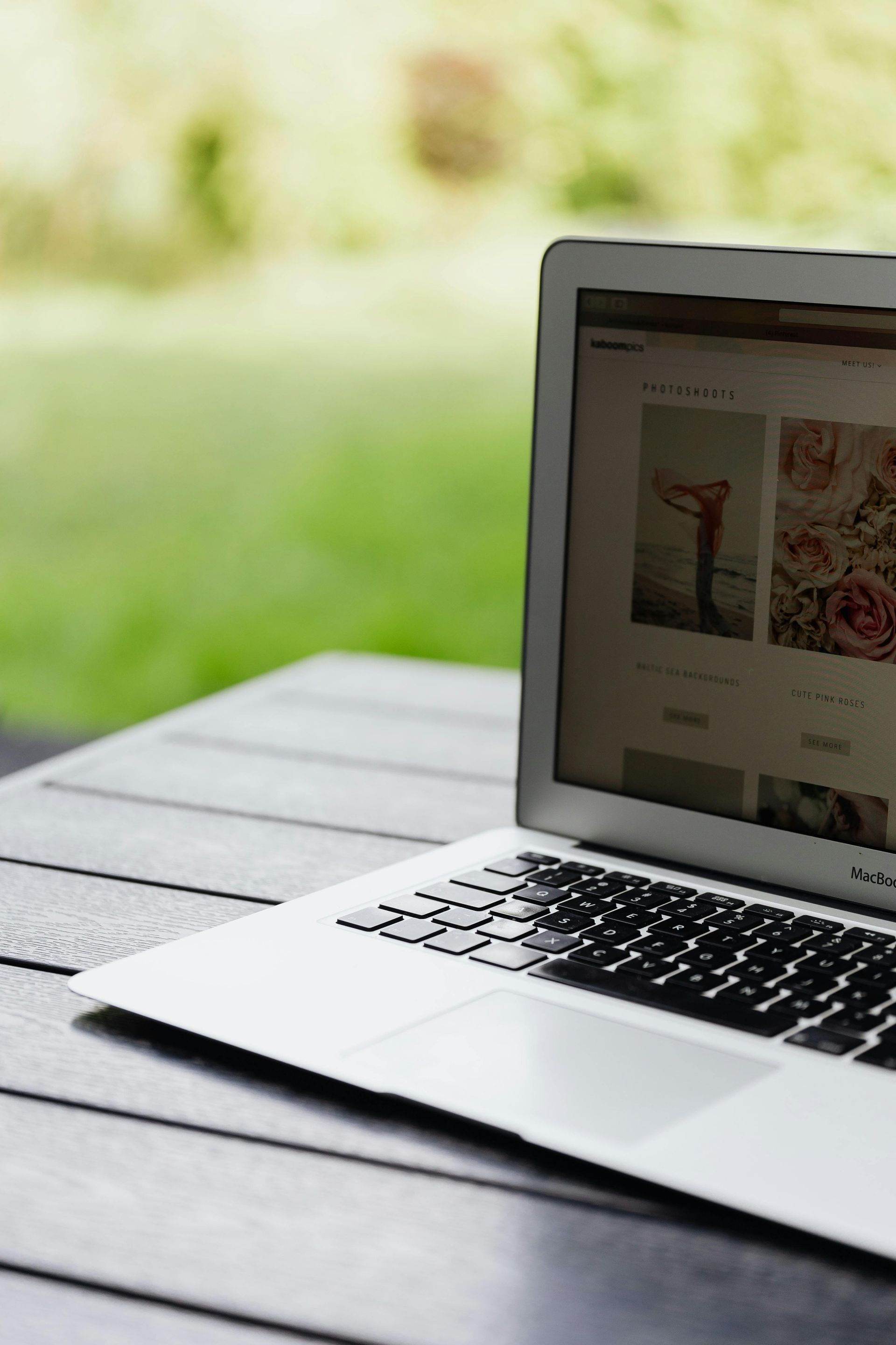 Laptop open on a wooden table outdoors, displaying a website with images of flowers. Green background.