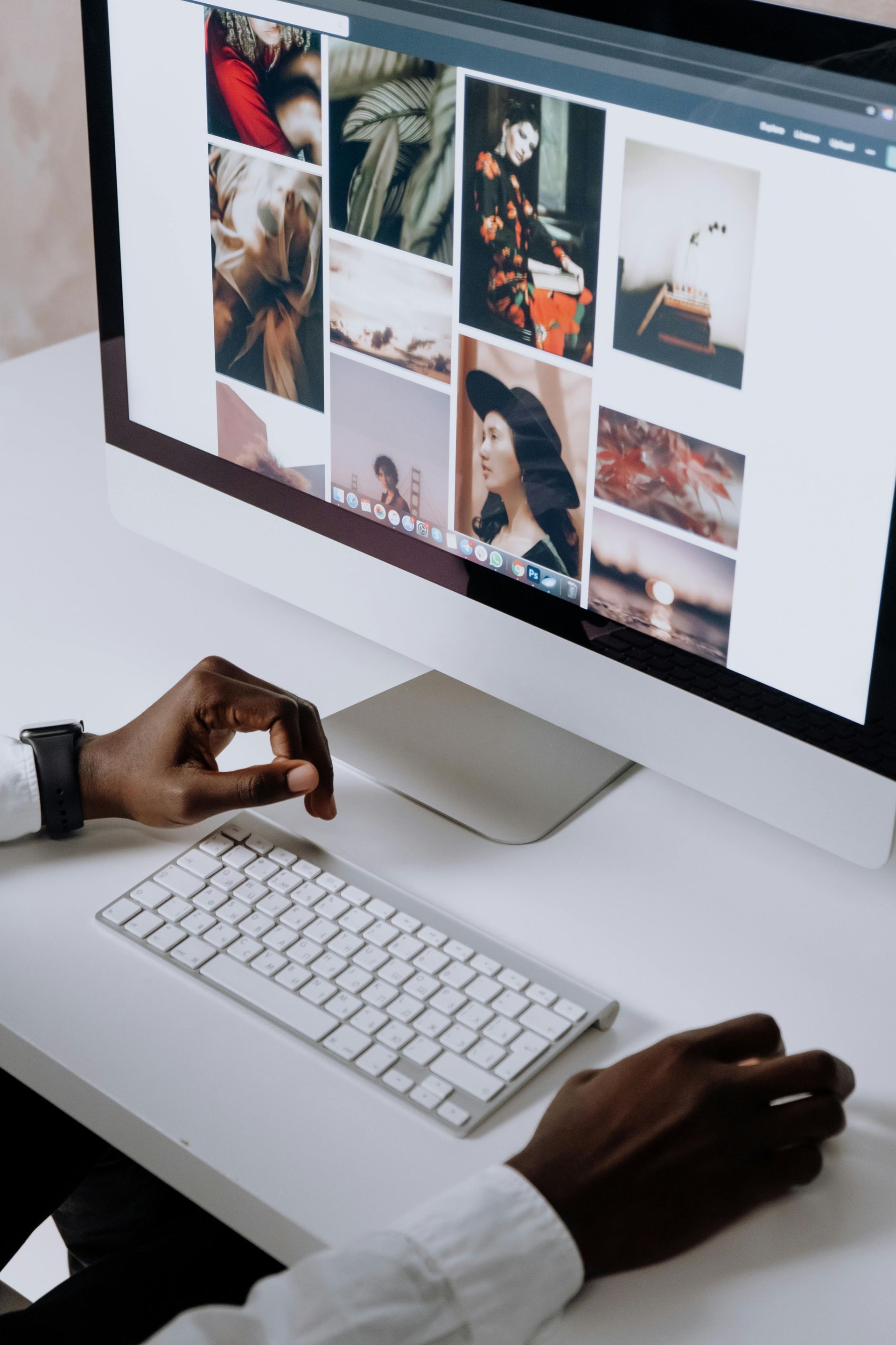 Person using computer, looking at a grid of images. Hands on keyboard and mouse, white desk setting.