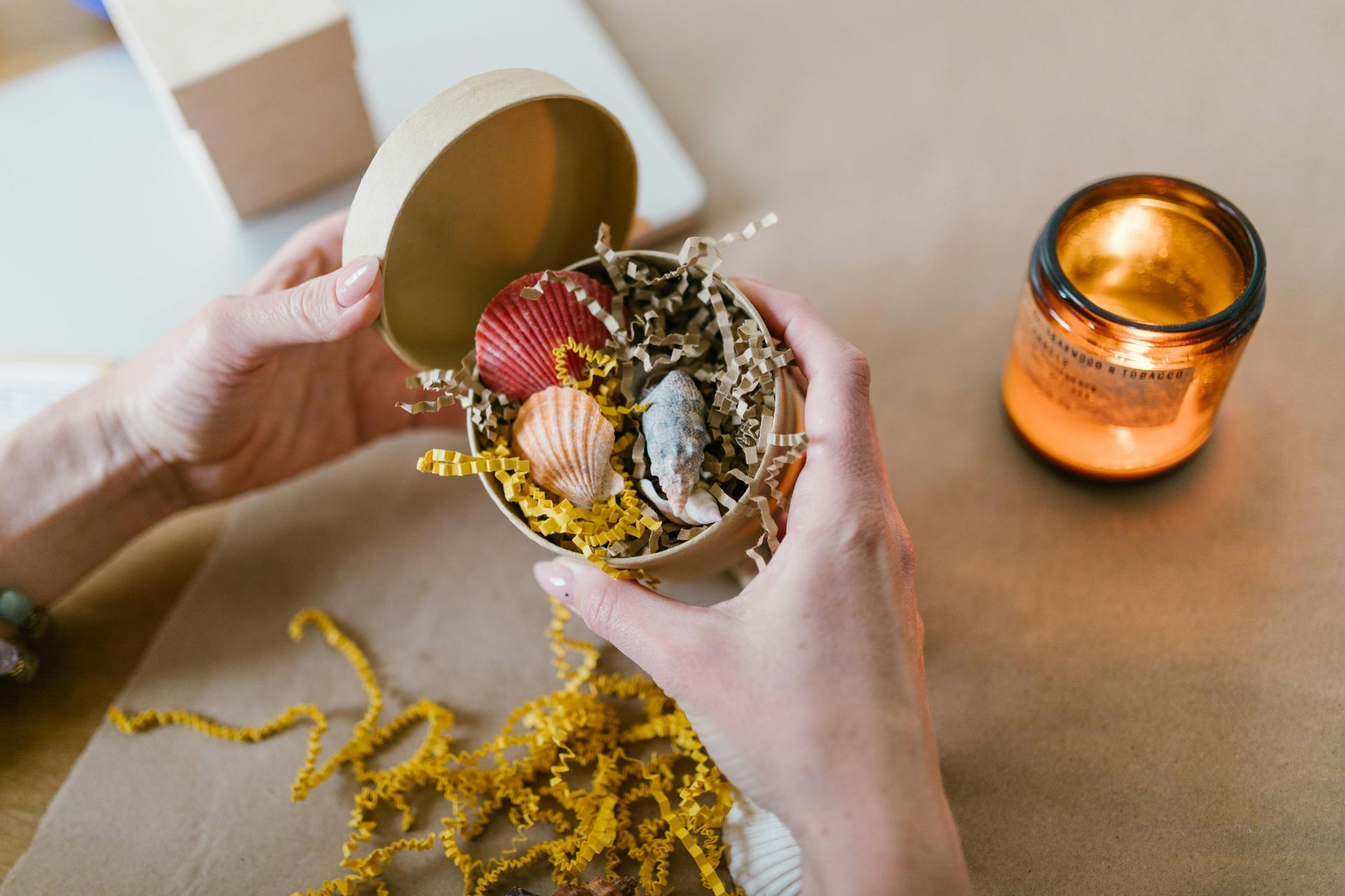Hands holding an open round gift box filled with seashells and shredded paper, with a lit candle beside it.