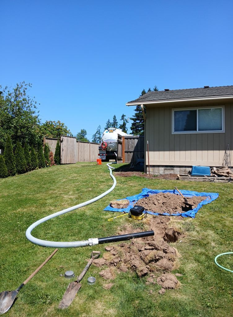 A septic tank being serviced in a grassy backyard. A truck and hose are connected to a buried system, with a pile of dirt nearby.