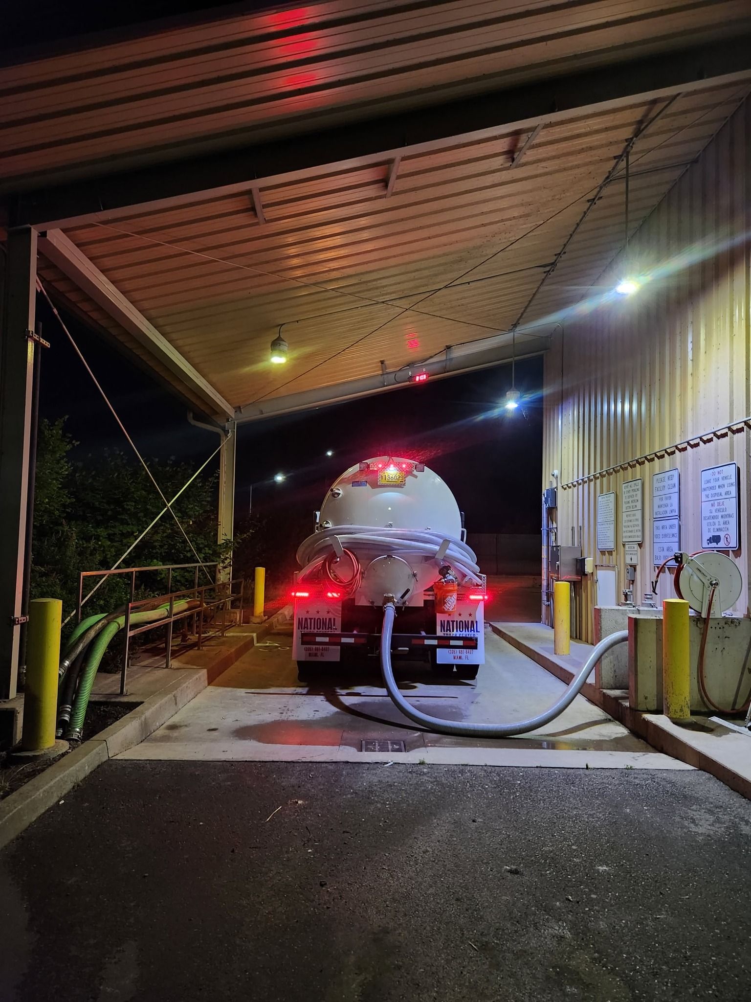 Tanker truck at a loading dock at night, with lights on. A hose connects to the truck.