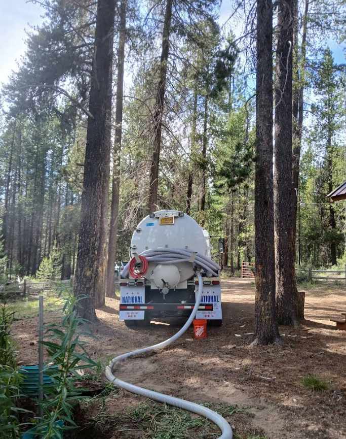 Tanker truck with white tank and gray hose, parked among tall trees, in a wooded area.