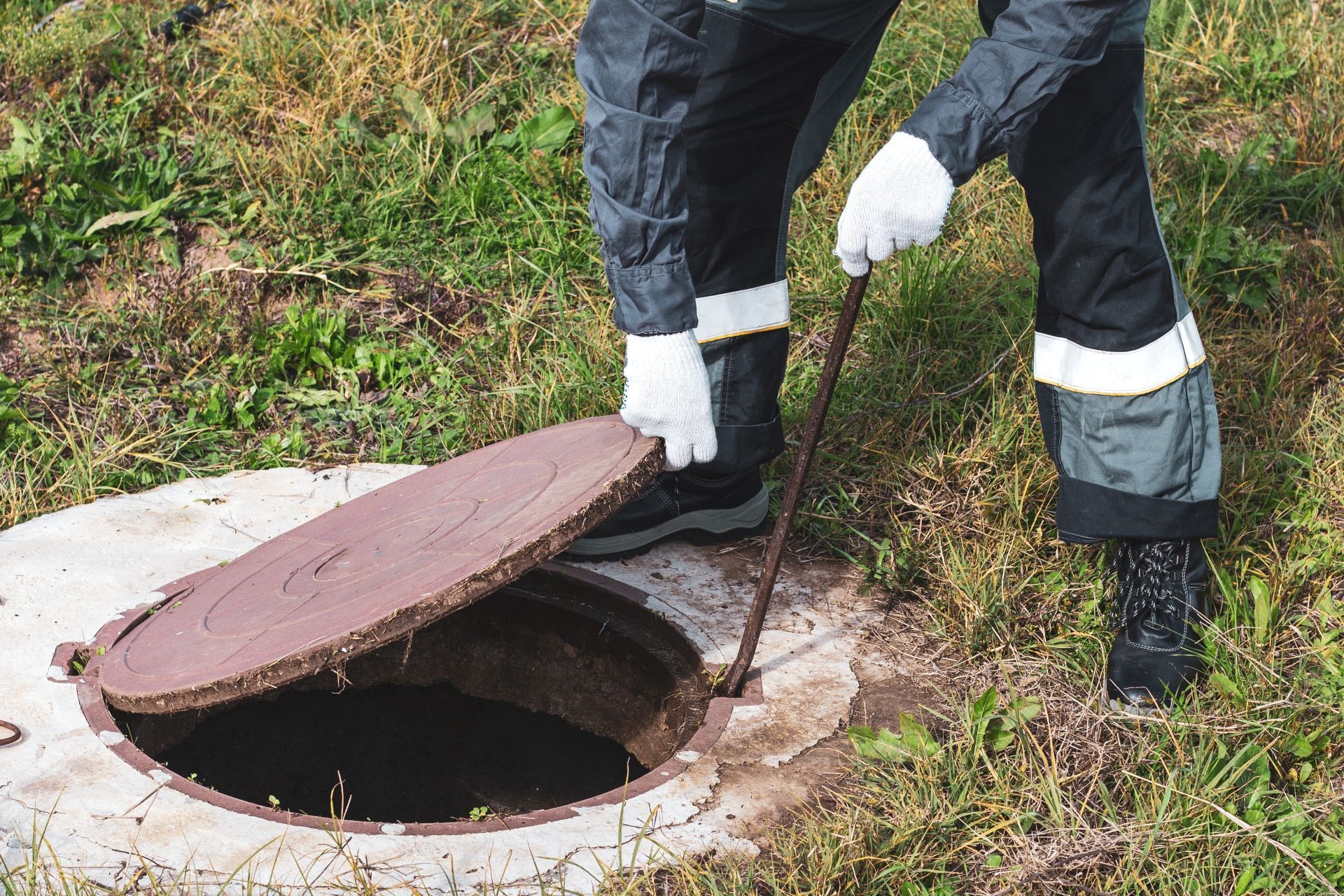 Person in protective gear opens a manhole cover in a grassy area.