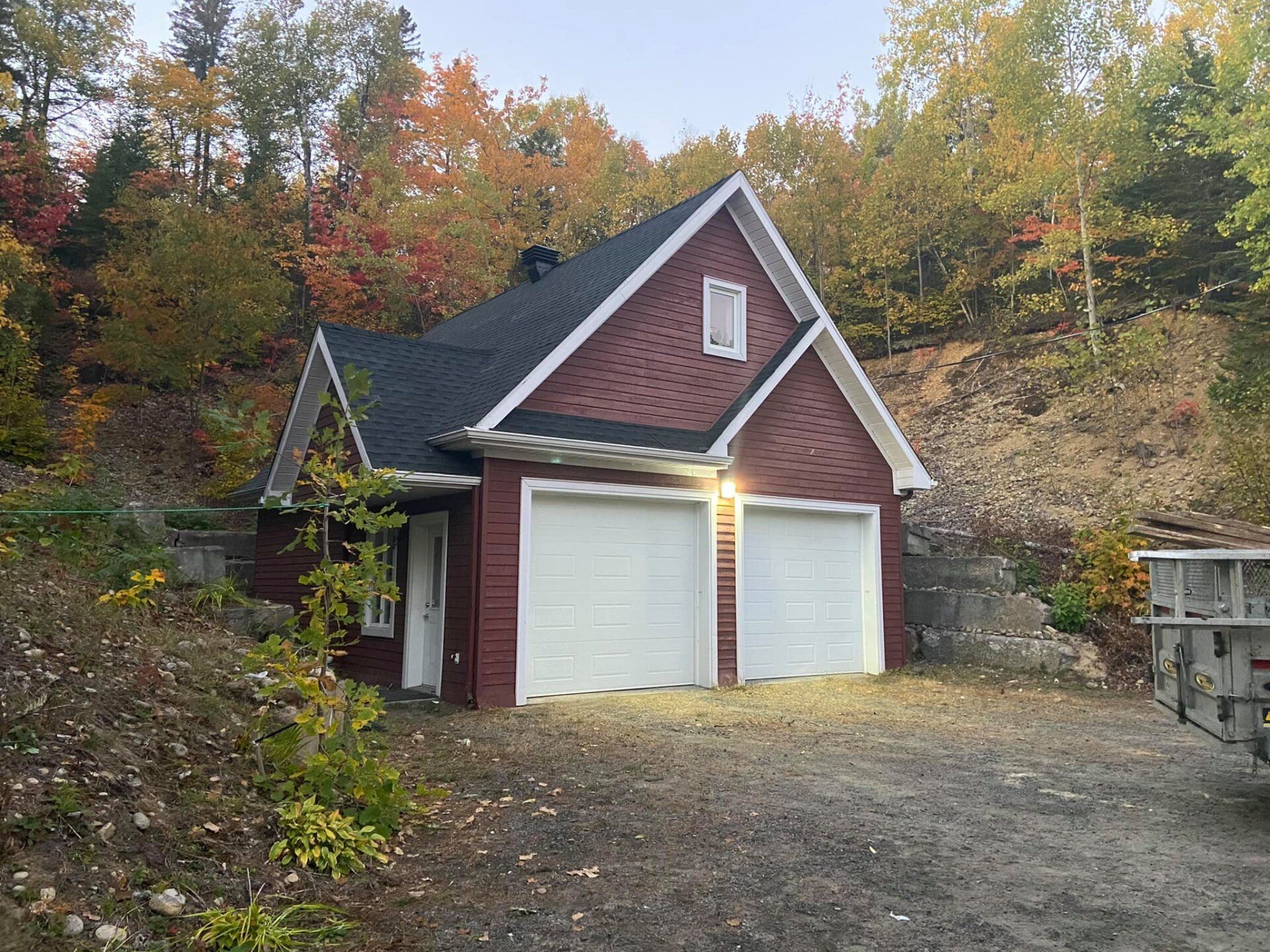 Garage rouge pour deux voitures, portes blanches, toit noir et arbres aux couleurs d'automne.