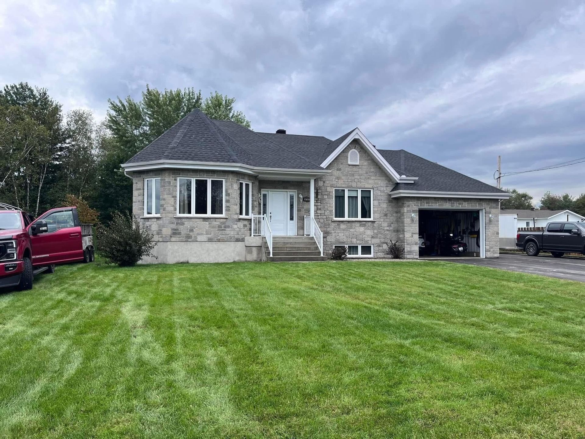 Maison en briques grises avec garage, porte blanche et pelouse verte sous un ciel nuageux.