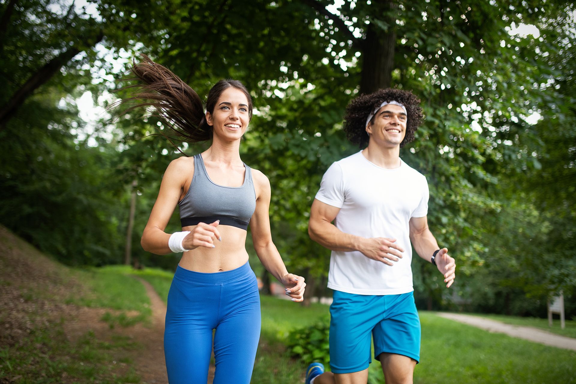 A smiling person in a grey sports bra and blue leggings and another in a white t-shirt and blue shorts jogging outdoors.