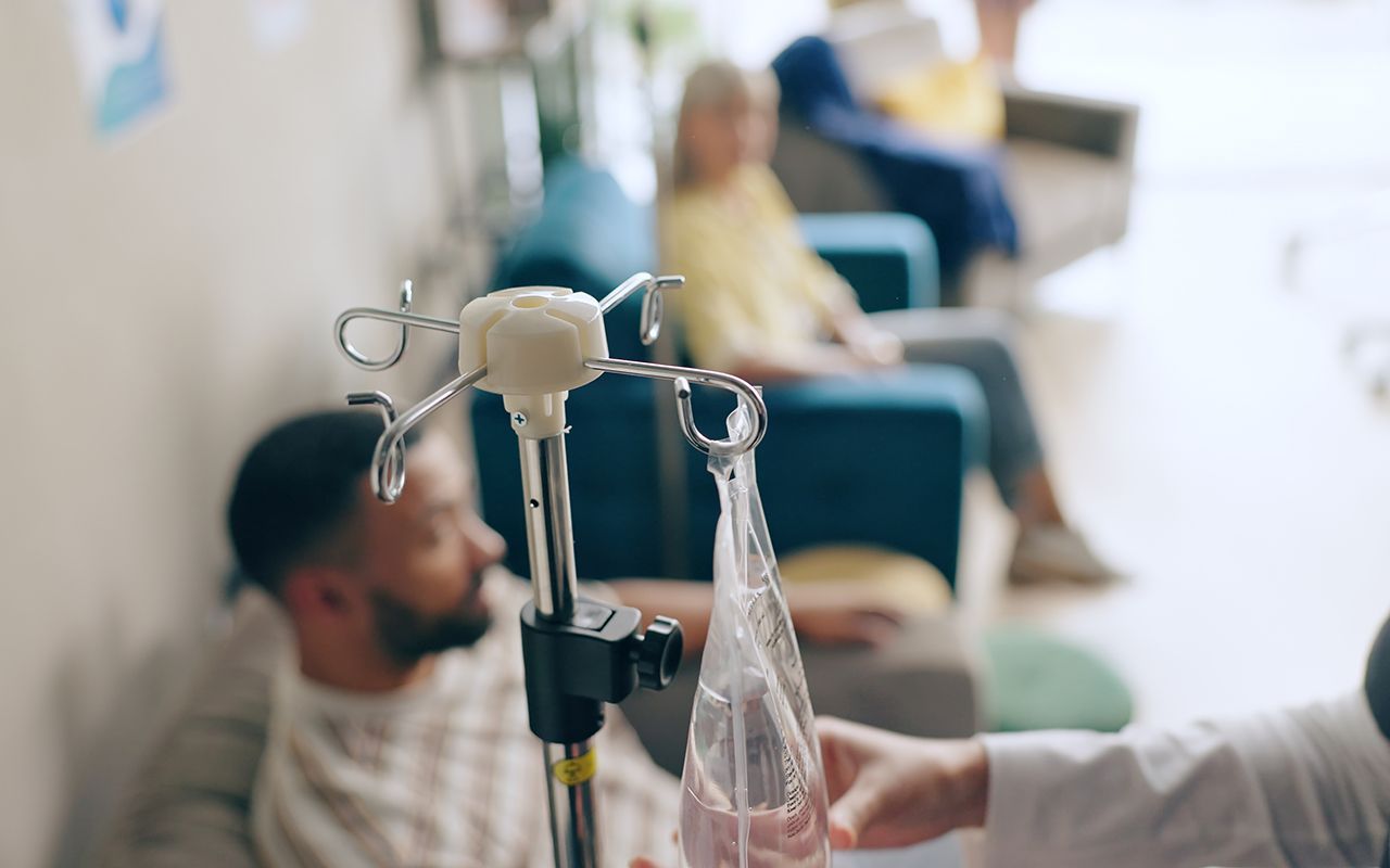 A medical professional adjusts an IV drip bag in a waiting area where patients are seated in the blurred background.