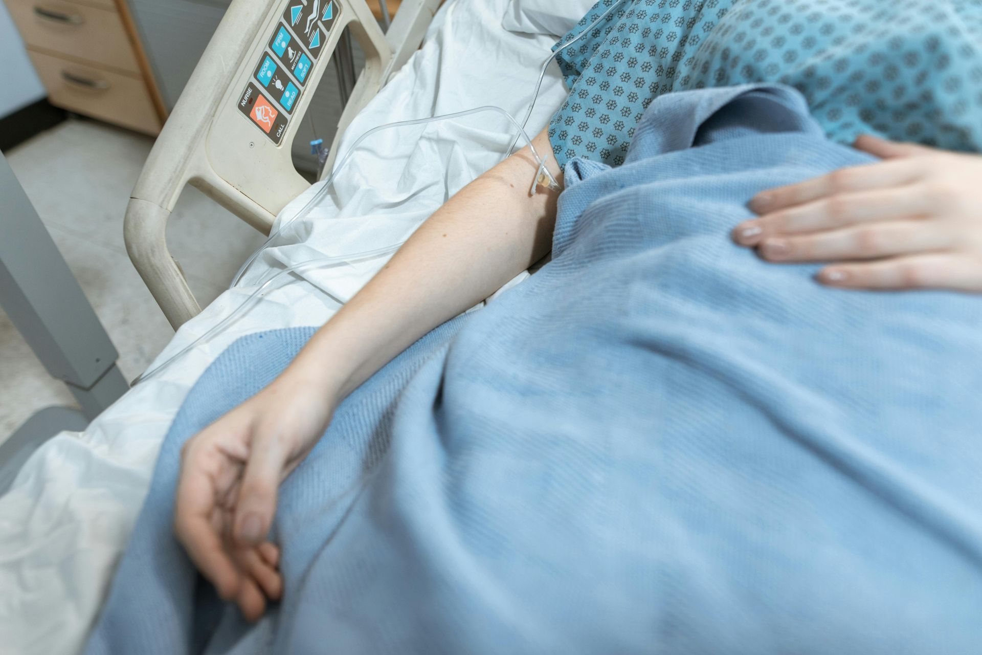 Person in hospital bed, arm with IV, covered by a blue blanket, resting on bed.
