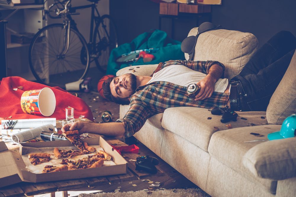 A man is laying on a couch in a messy living room.