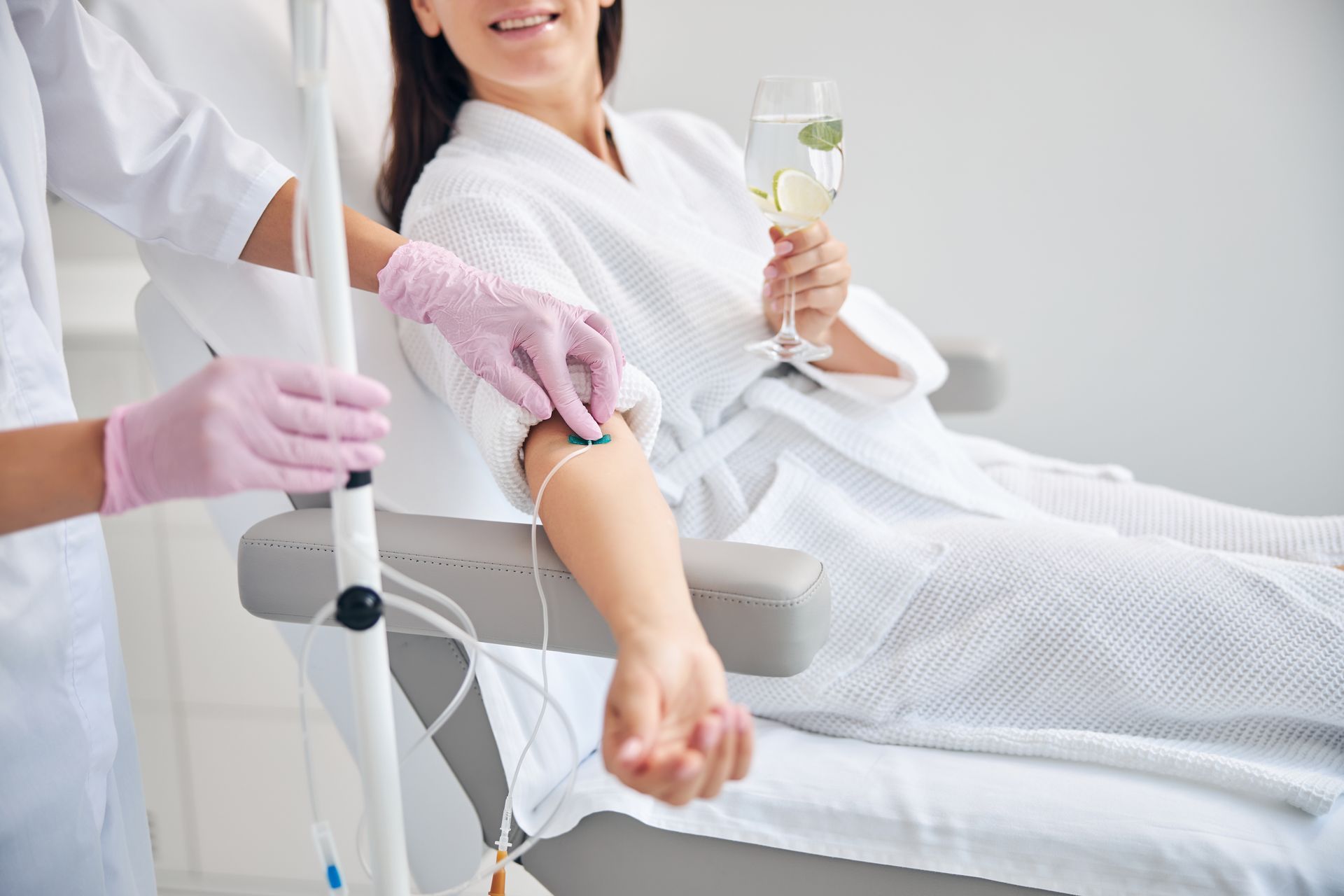 A healthcare worker in gloves attaches an IV line to a person in a bathrobe holding a glass of infused water.