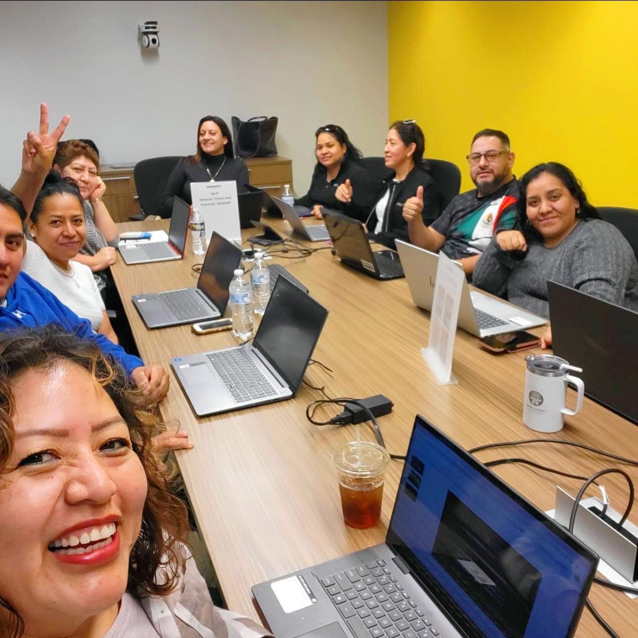 People at a long table with laptops in a brightly lit room, looking at the front.
