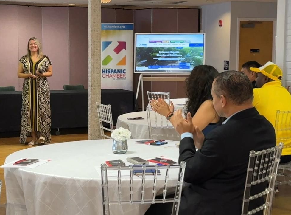 Woman presenting at a table to seated audience, with screen and banner in a room.