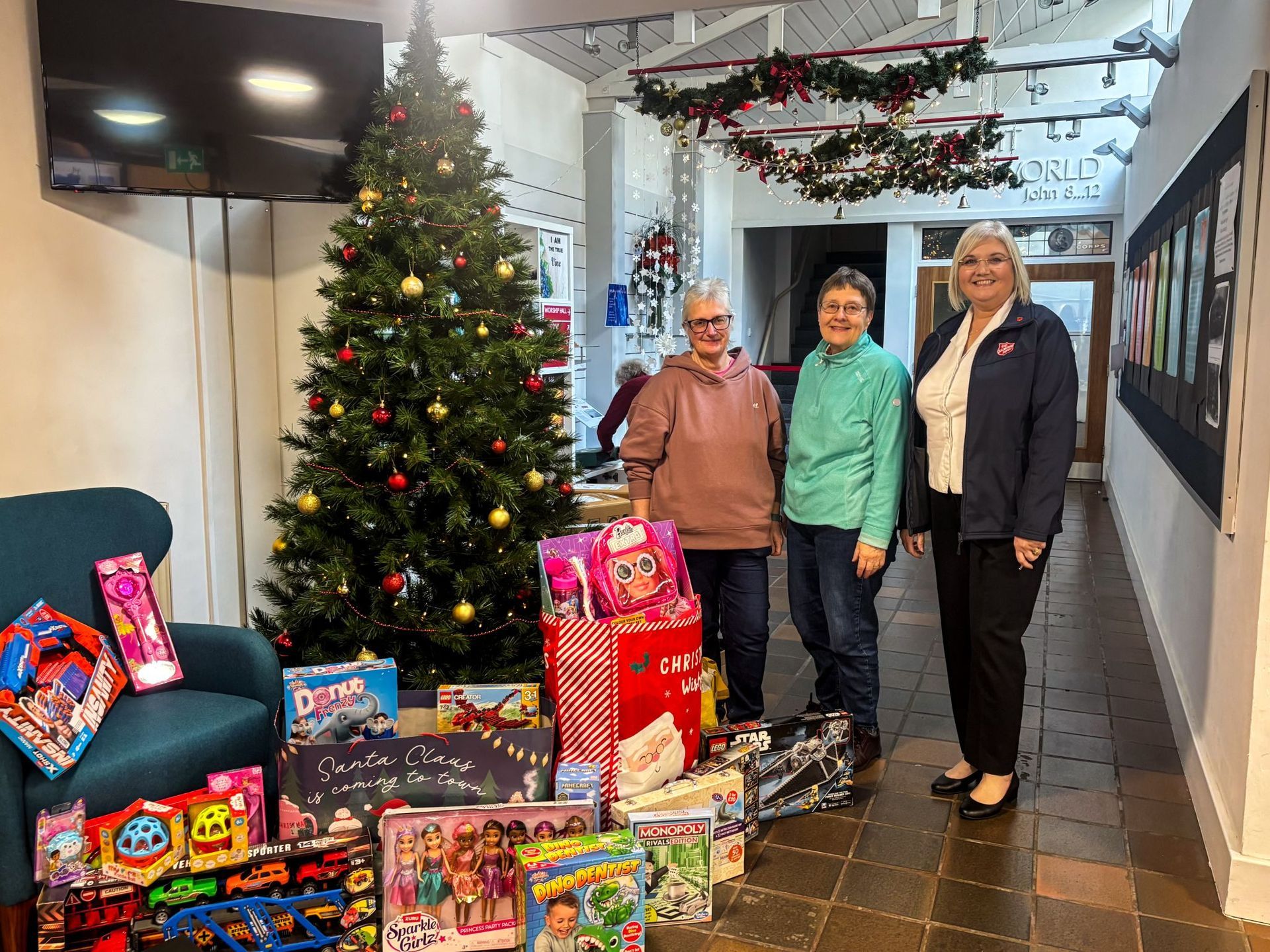 Three people are standing in front of a christmas tree surrounded by gifts.