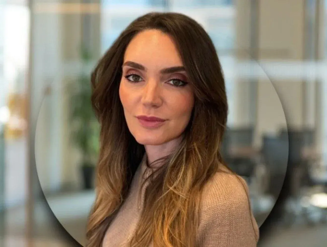 A person with long brown hair, wearing a neutral-colored top, looking directly at the camera in a professional office.