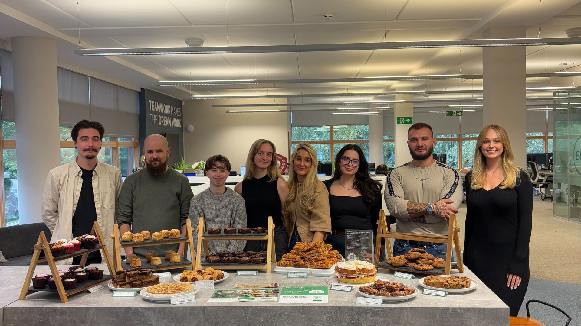 People stand behind a table of baked goods, smiling. Office setting.