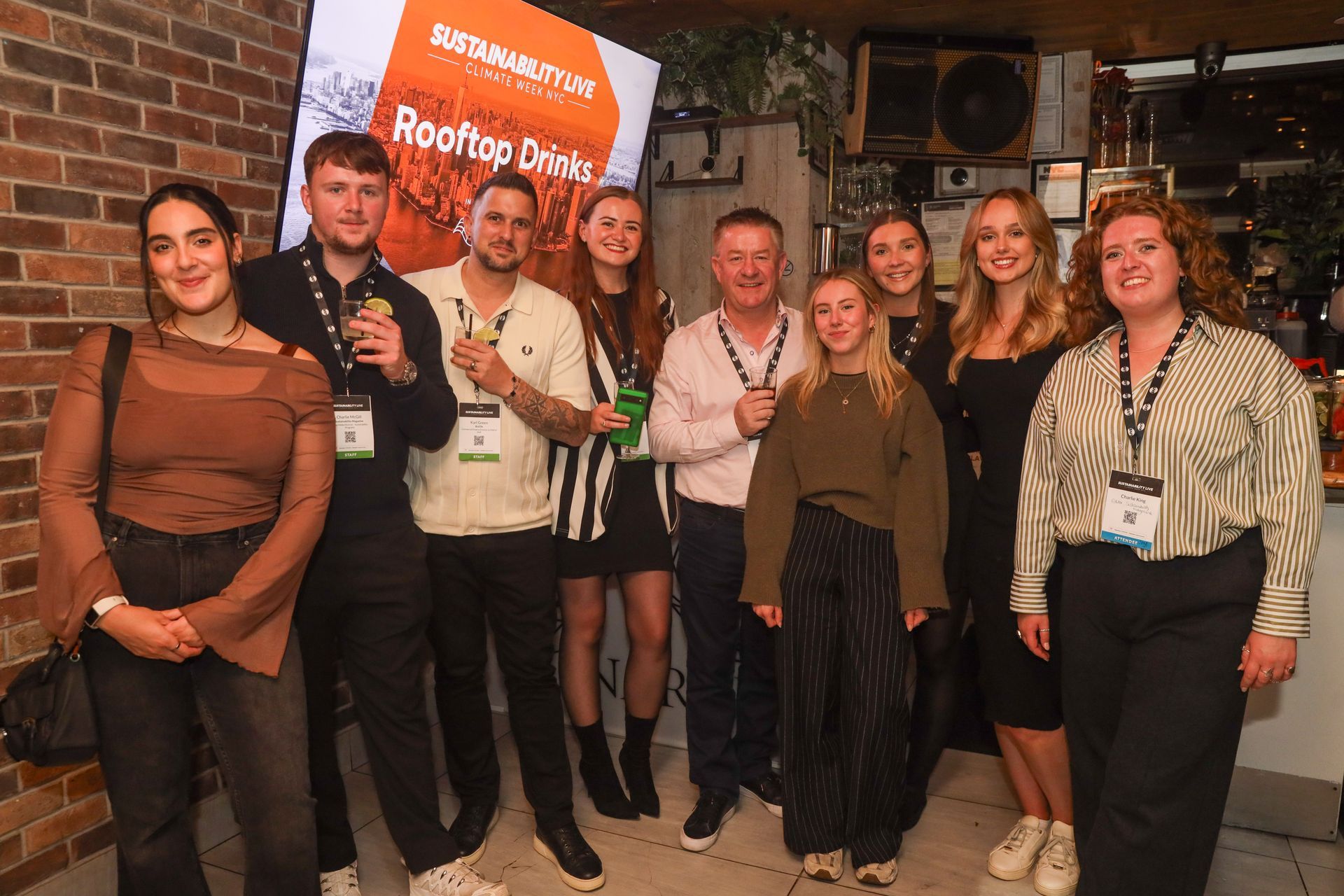 Group of people smiling, indoors. Some hold drinks, wearing lanyards. Background brick wall and promotional material.