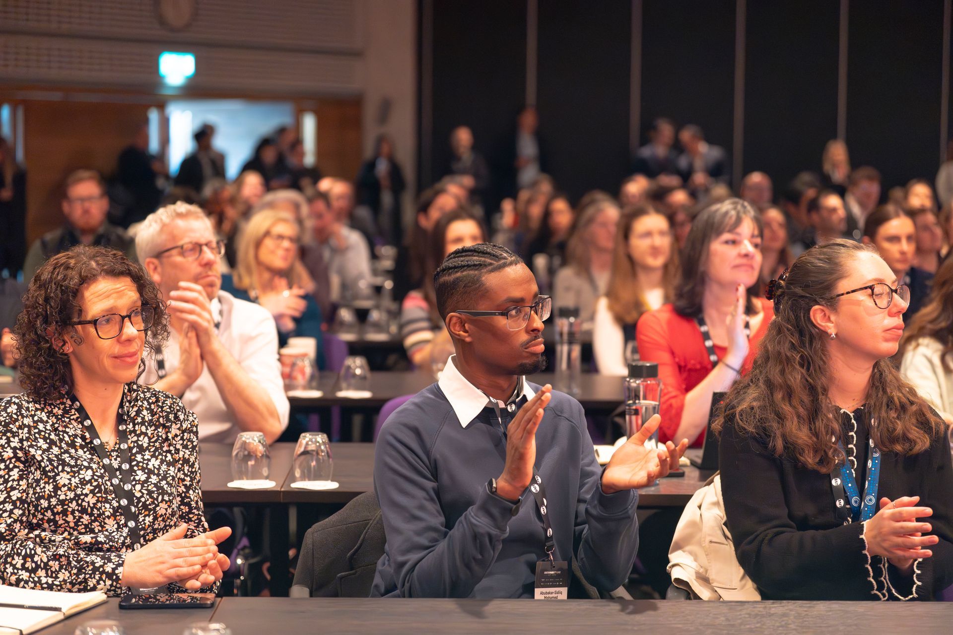 A group of people are sitting at tables in a room clapping their hands during Sustainability LIVE.