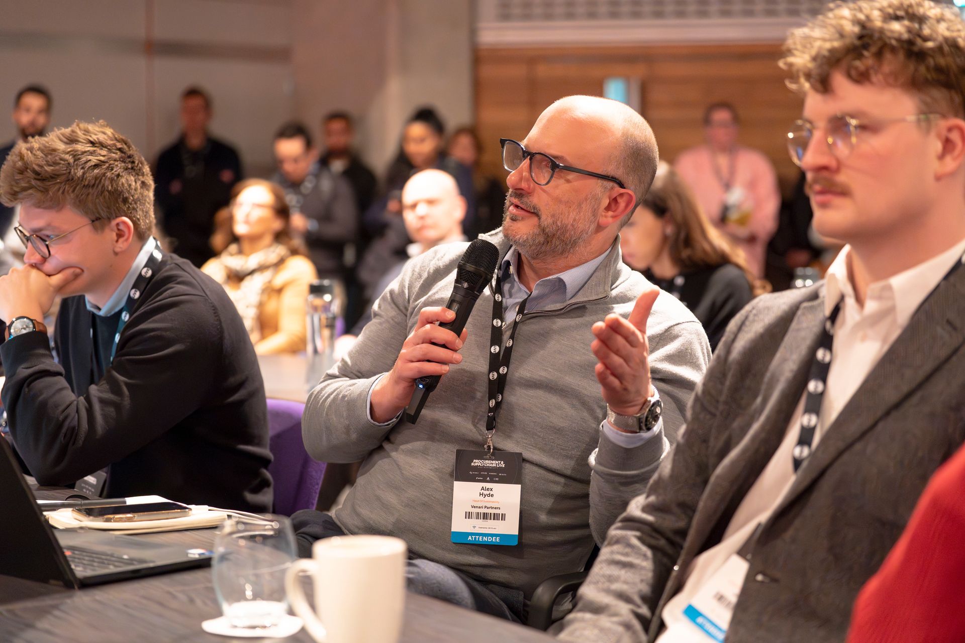 A man is sitting at a table holding a microphone and talking to a group of people.