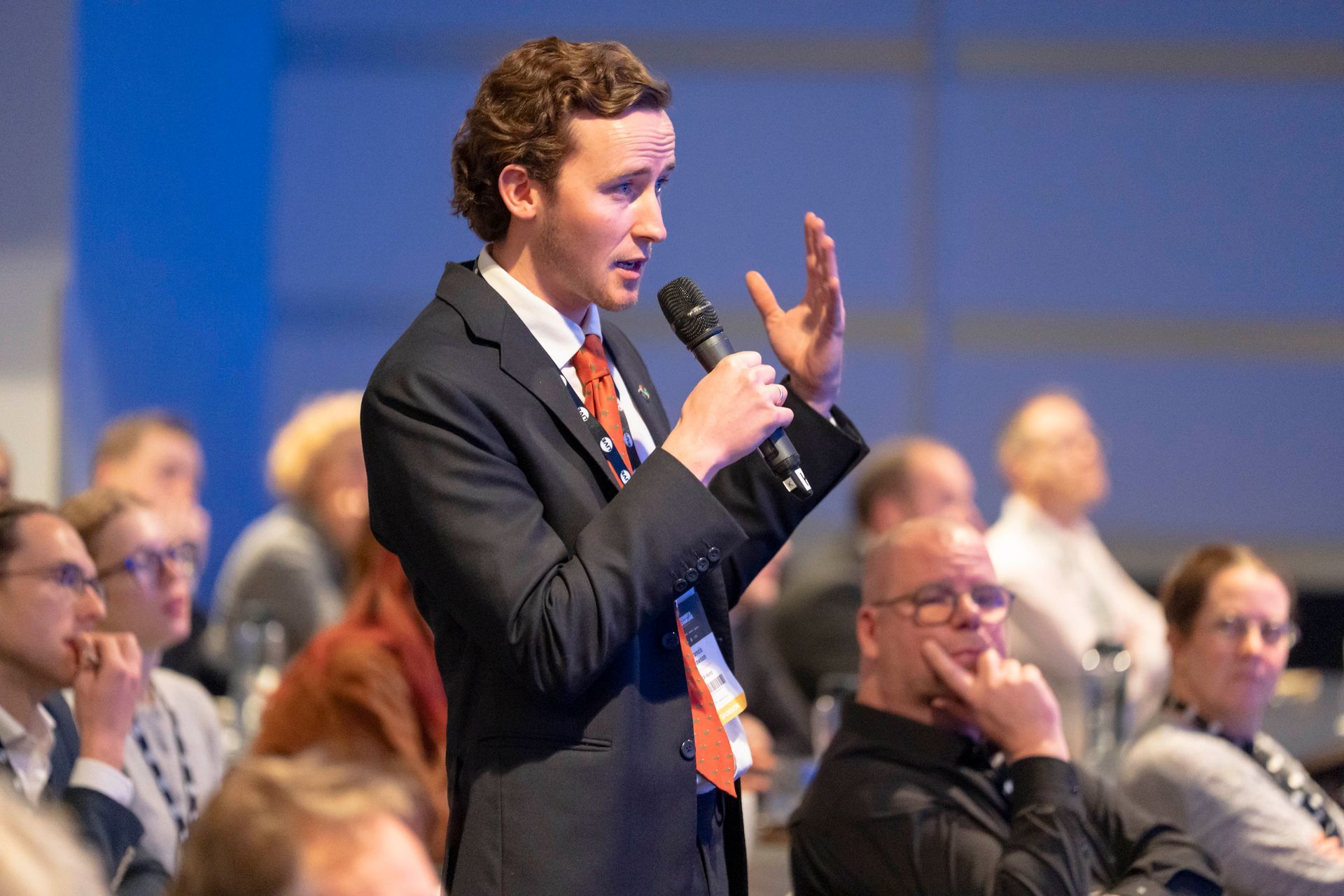 A man in a suit and tie is holding a microphone and talking to a group of people.