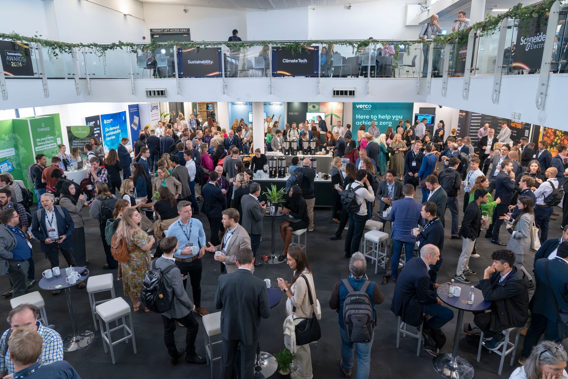 A large event with many people mingling in a bright, open venue. Groups of attendees chat and network near tables and bars.