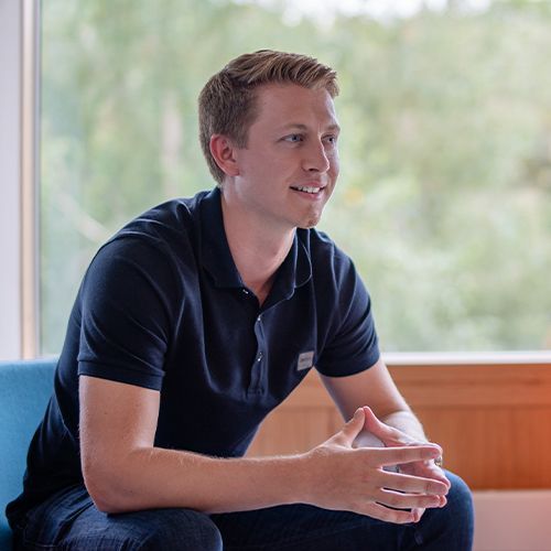 A man in a black shirt is sitting on a blue couch in front of a window.