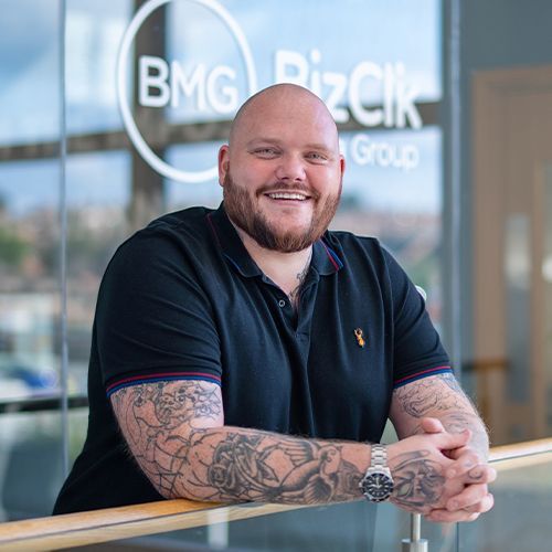 A bald man with a beard and tattoos is leaning on a railing.