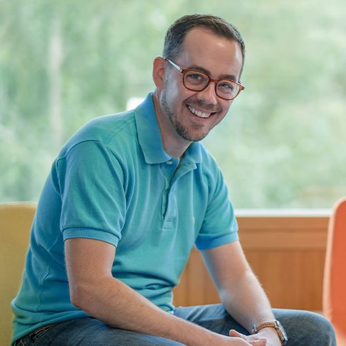 A man wearing glasses and a blue shirt is sitting on a chair and smiling.