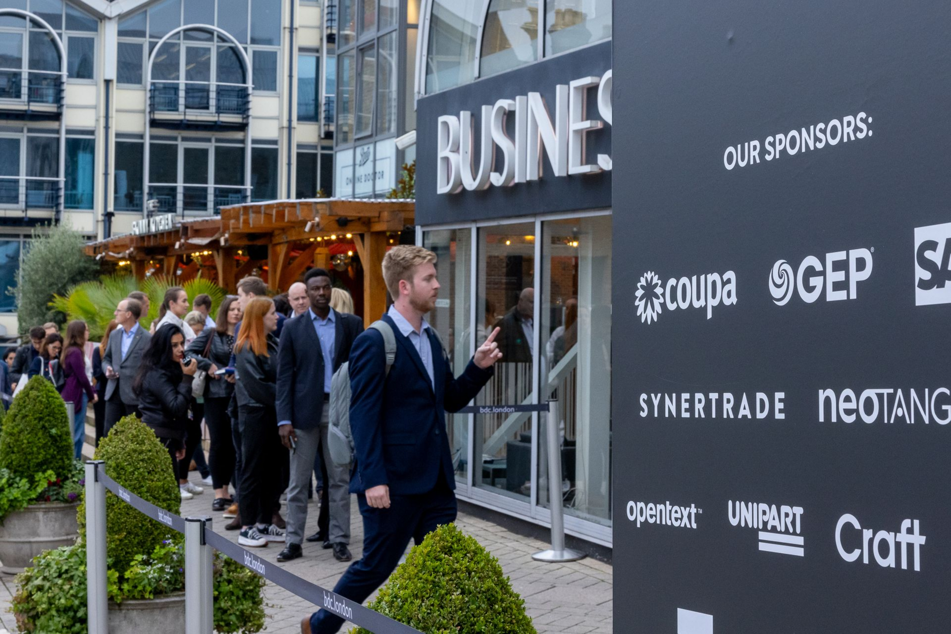 A large event with many people mingling in a bright, open venue. Groups of attendees chat and network near tables and bars.