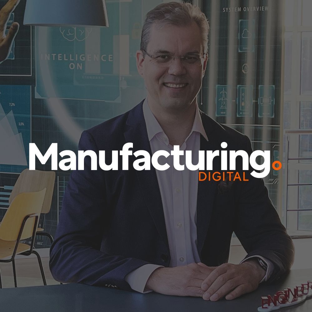 A man in a suit is sitting at a table in front of a manufacturing digital logo.