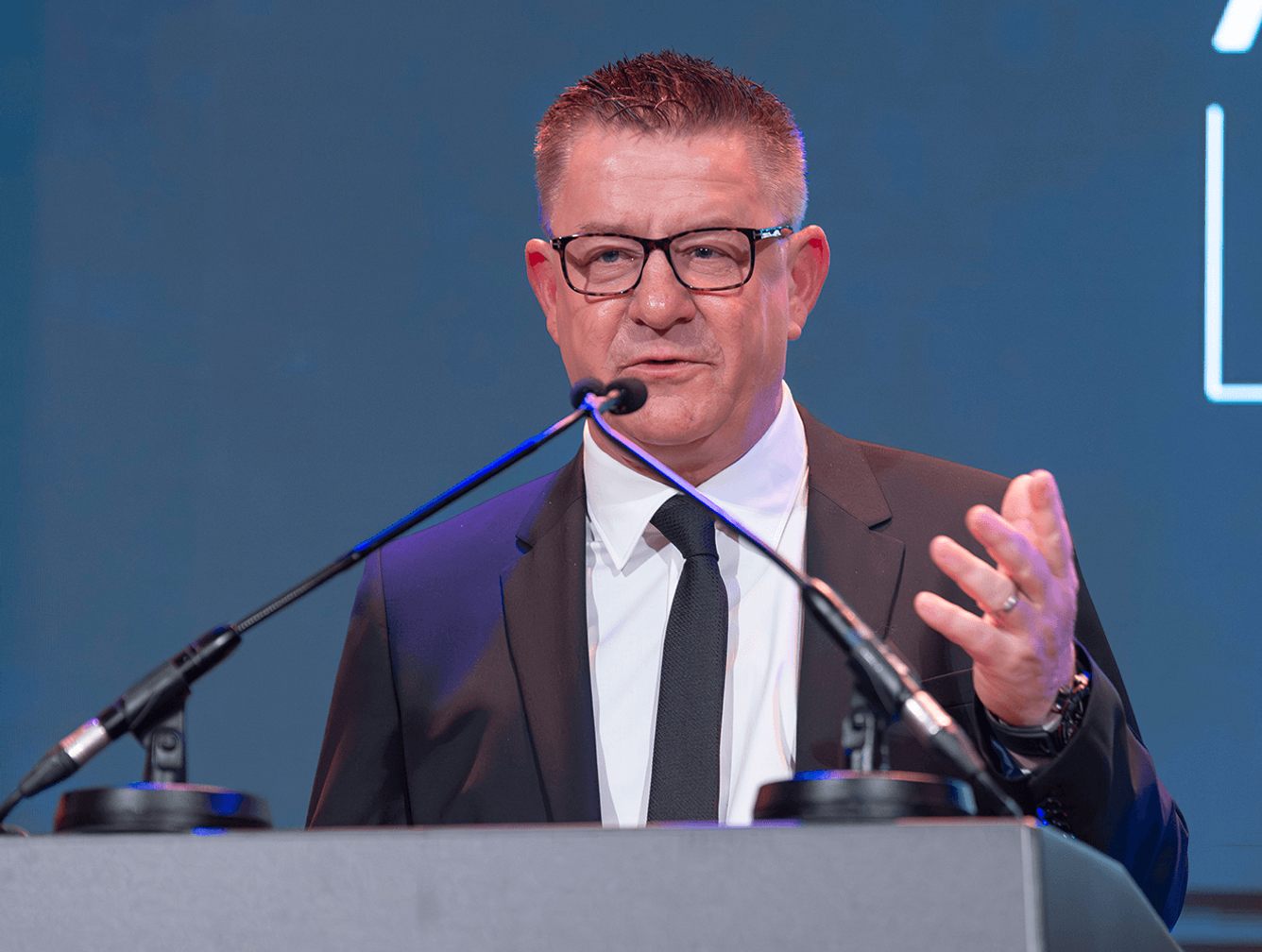 A man in a suit and tie speaks at a podium with two microphones against a blue backdrop.