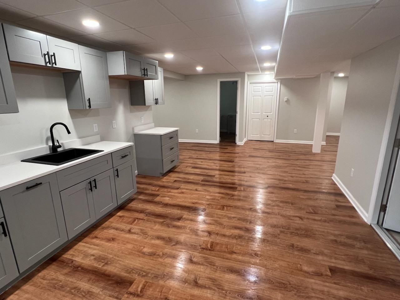 A kitchen in a basement with hardwood floors and white cabinets.