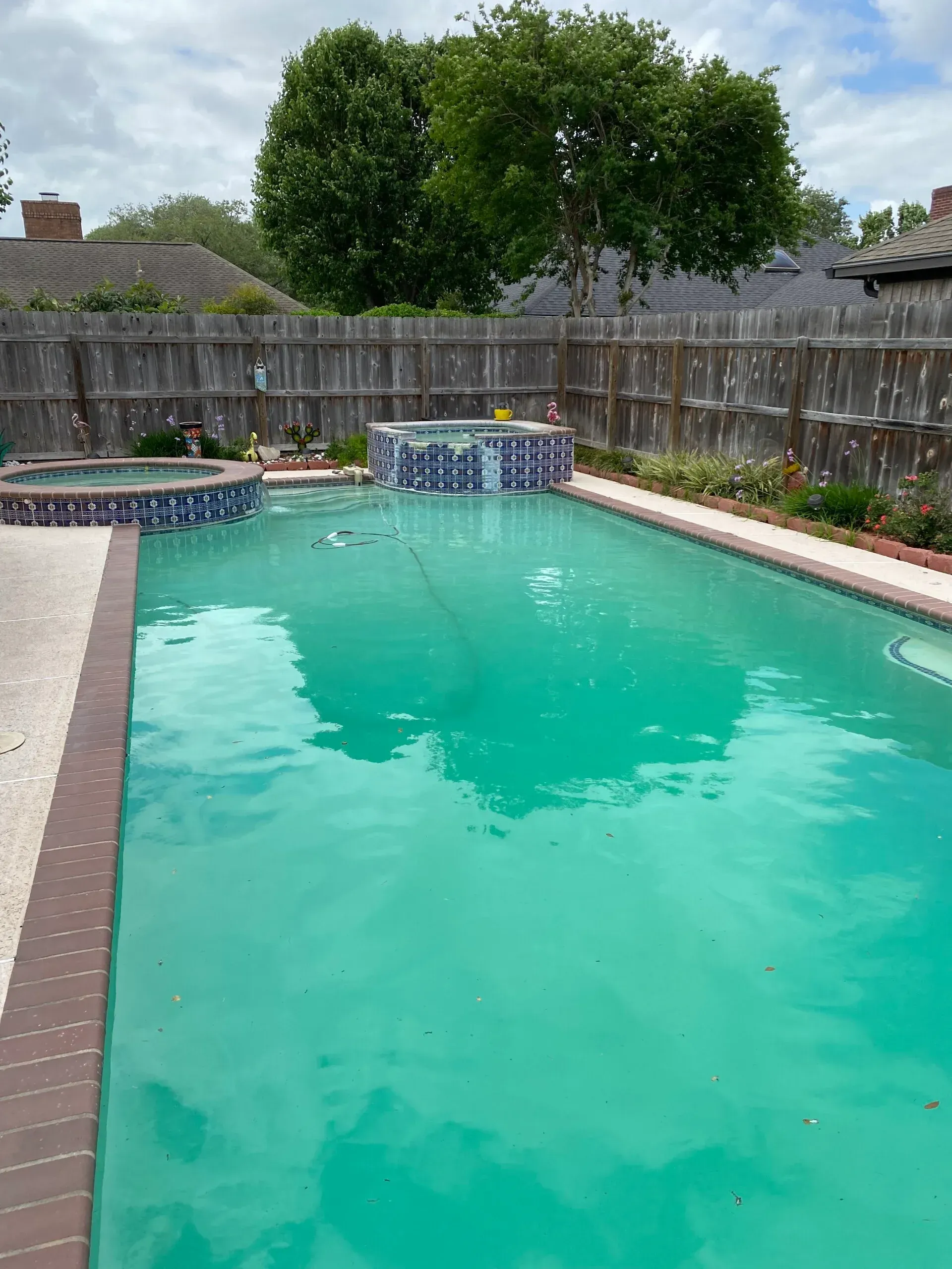Swimming pool with turquoise water, a hot tub, and a decorative waterfall feature against a wooden fence.