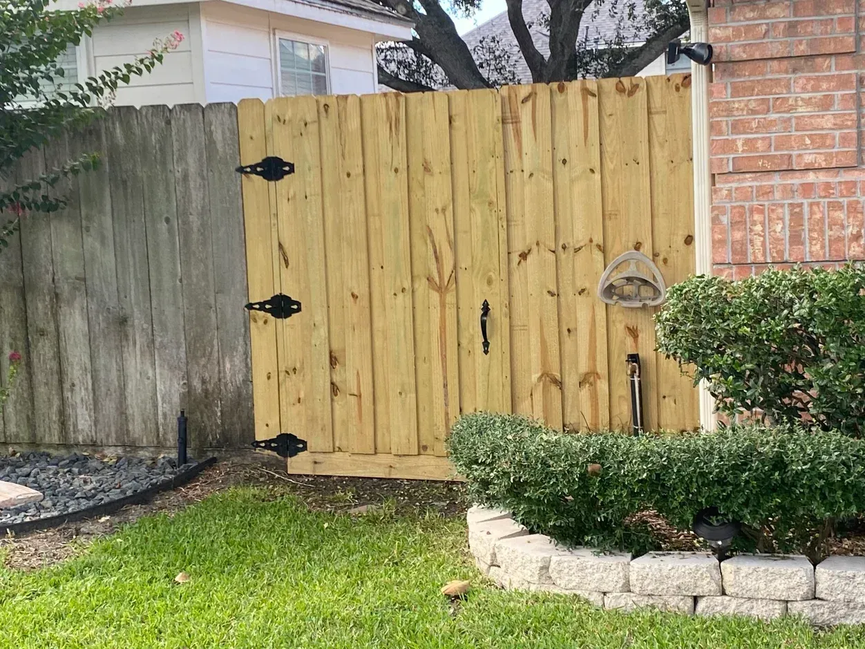 A wooden gate in a yard, connected to an older wooden fence and a brick wall, with greenery.