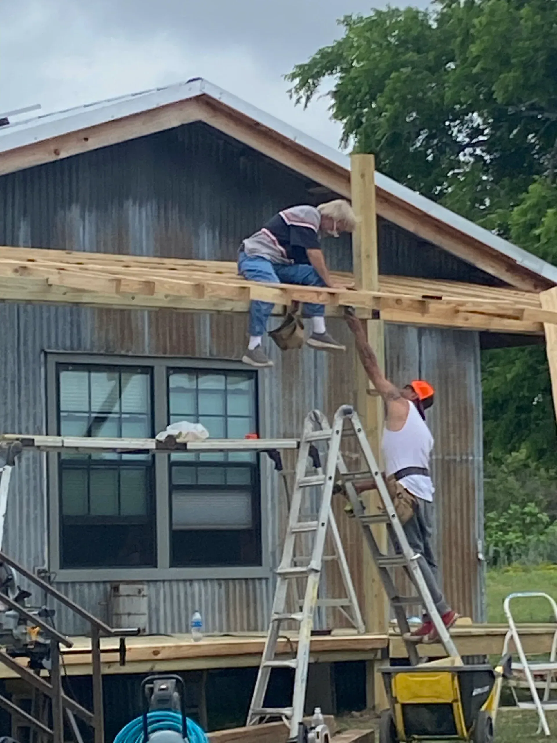 Two people building a wooden structure attached to a metal-sided building. One on a ladder, one on the roof.