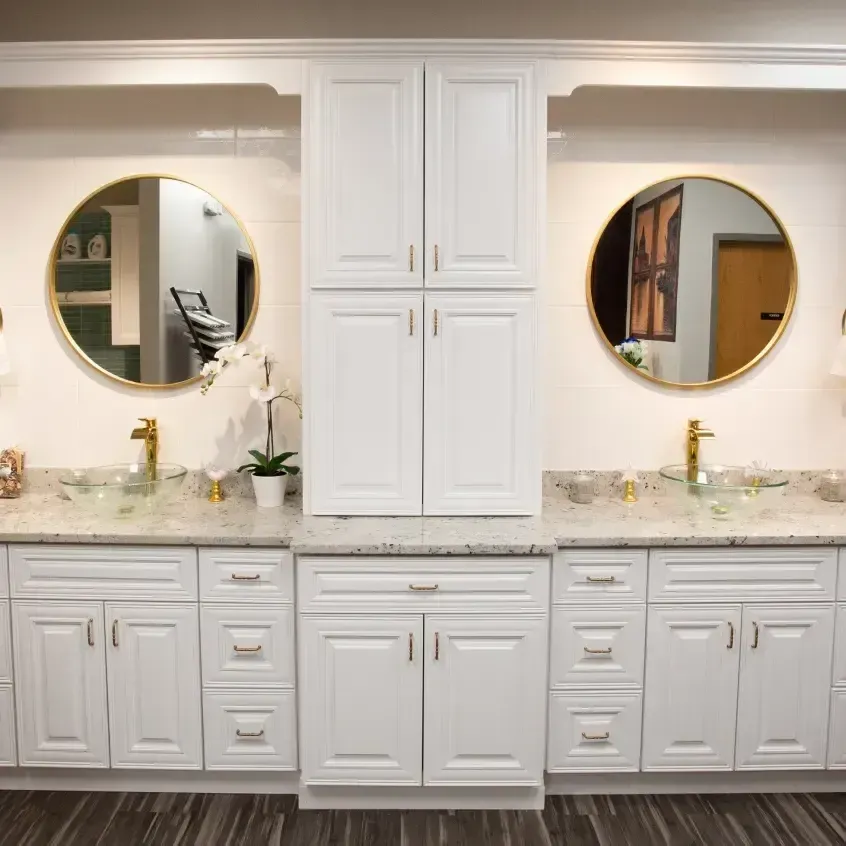 A modern white bathroom with two sinks, gold fixtures, round mirrors, and a central storage cabinet.