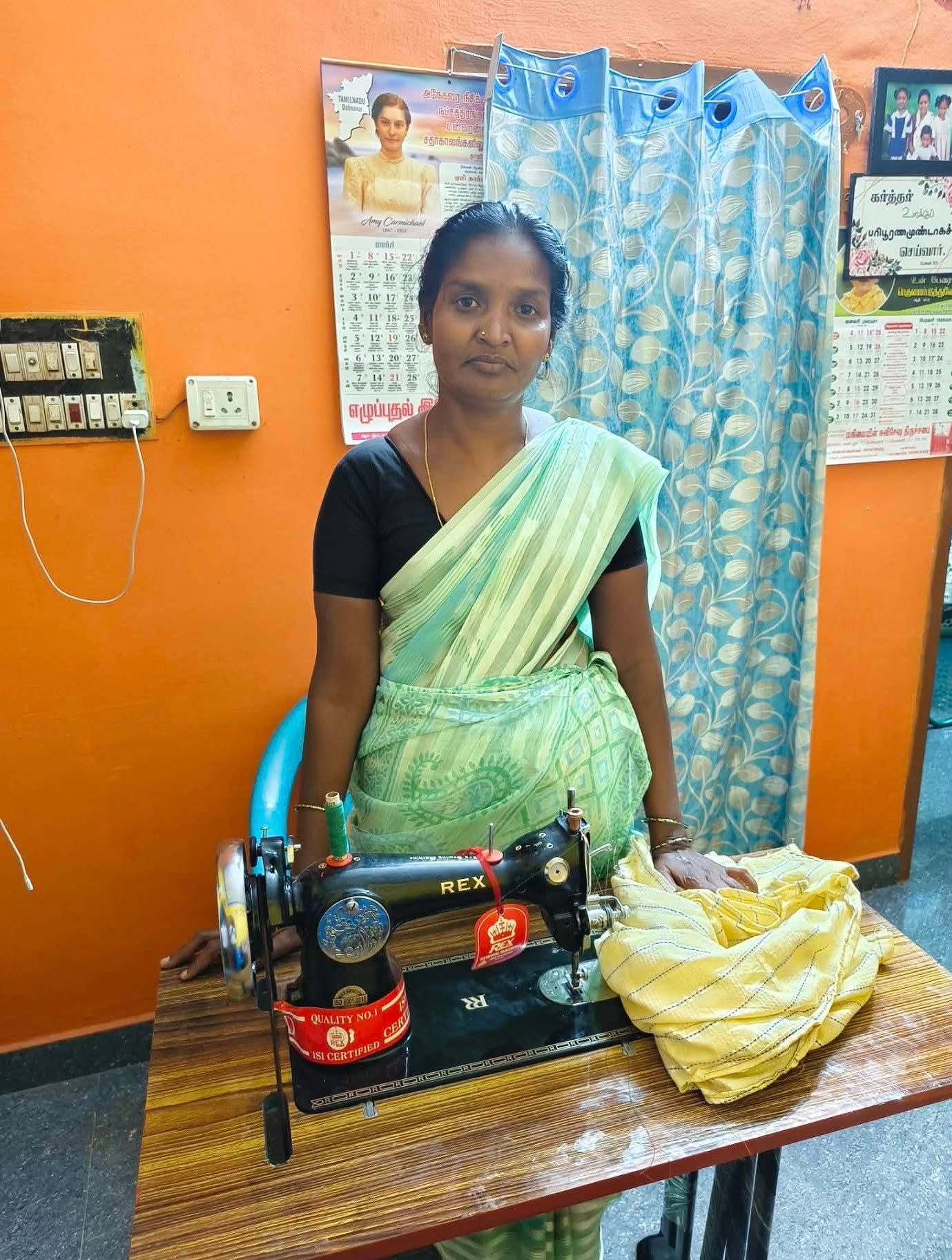 A person wearing a light green and white saree sits at a sewing machine with a pile of yellow fabric on the table.