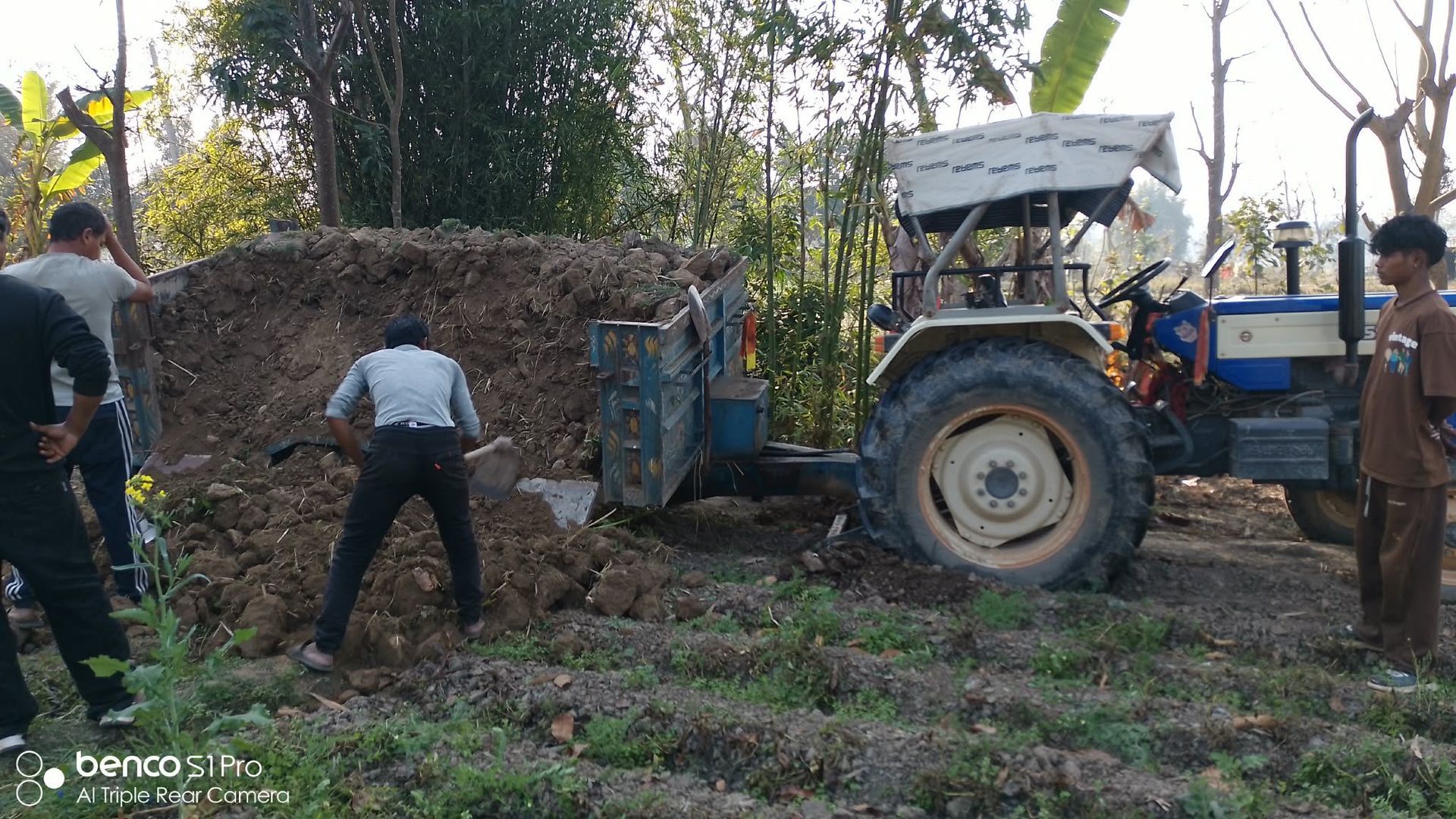 Three people working near a tractor-drawn trailer that has dumped a large pile of dirt in a rural field.